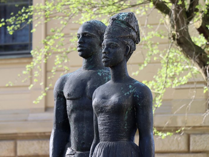Bronzestatue von zwei eng stehenden Personen, eine mit langem Gewand und Kopfbedeckung, die andere mit kurzem Gewand, die sich gegenseitig umarmen oder stützen.