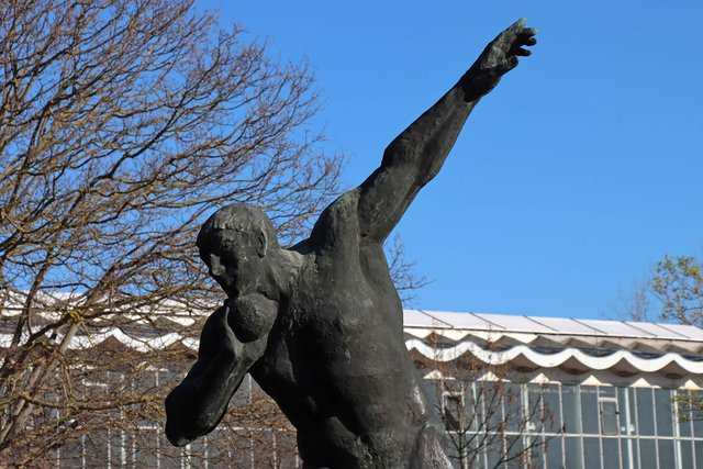 Bronzestatue eines nackten Menschen, der mit einem Arm nach hinten gestreckt und dem anderen Arm angewinkelt in der Luft schwebend dargestellt wird, vor klarem Himmel und winterlicher Baumkulisse.