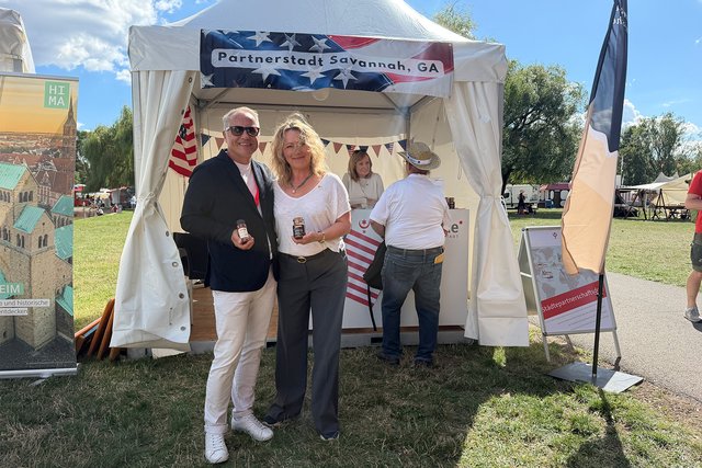 Zwei Personen stehen vor einem Informationsstand mit dem Schild „Patrioten für Amerika“. Sie halten jeweils eine kleine Flagge der USA und lächeln. Der Stand ist mit amerikanischen Symbolen geschmückt.
