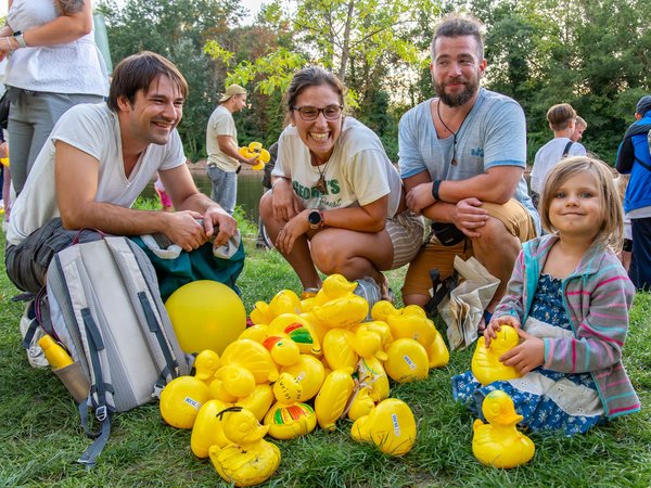 Eine Gruppe von vier Personen, darunter ein Kind, sitzt auf dem Gras und lächelt. Vor ihnen liegen viele gelbe Enten aus Plastik verstreut.