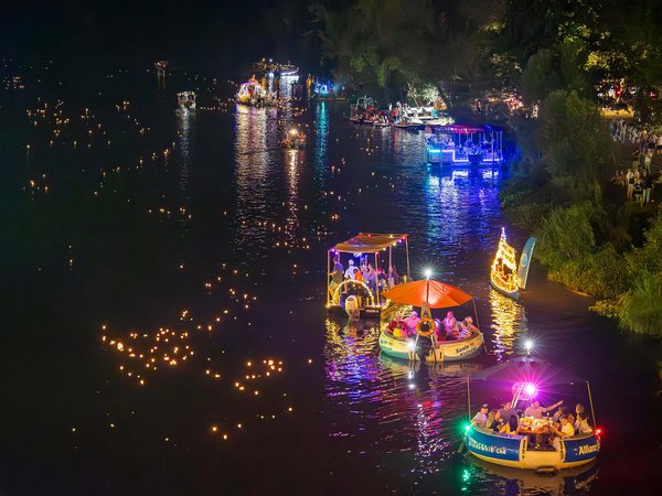 Nachts beleuchtetes Boot auf einem Fluss mit farbigen Lichtern und Dekorationen, umgeben von anderen festlich geschmückten Booten, die ebenfalls illuminiert sind.