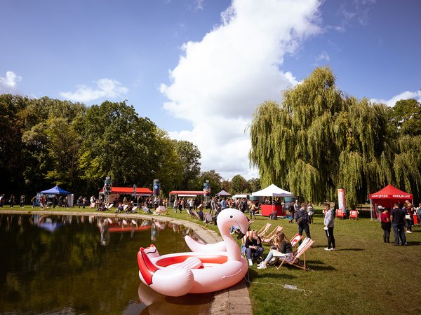 Großer Enten-Gummiboot mit rotem Schnabel und orangefarbenen Füßen liegt auf einem See. Im Hintergrund ein Fest mit Zelten, Menschen und Bäumen bei sonnigem Wetter.
