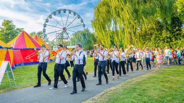 Eine Gruppe von uniformierten Personen, vermutlich Schüler oder Kadetten, marschiert im Gleichschritt auf einem Rasenweg. Im Hintergrund sind ein buntes Zelt, ein Riesenrad und grüne Bäume zu sehen.