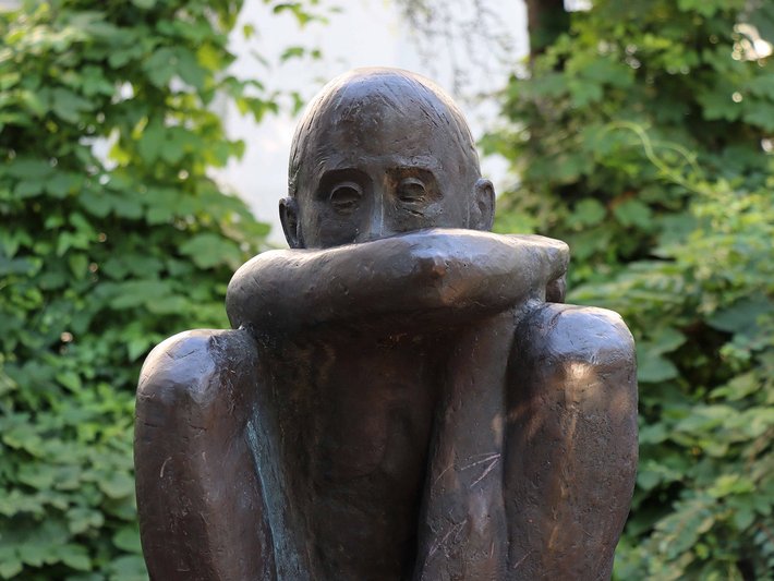Bronzestatue eines sitzenden Menschen, der die Hände vor dem Gesicht hält, in nachdenklicher oder trauriger Haltung.