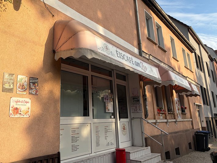 Fassade eines kleinen Cafés oder Restaurants mit dem Namen „Caffè Lario“ und einer hellen Markise. Im Schaufenster sind Speisekarten und Poster mit Getränke- und Essensbildern zu sehen.