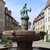 Bronzestatue einesreitenden Mannes auf einem Brunnen mit Wasserschale, umgeben von zwei weiteren Figuren, die auf einem steinernen Becken stehen. Historische Altstadtkulisse mit alten Gebäuden und Efeu bewachsenen Wänden.