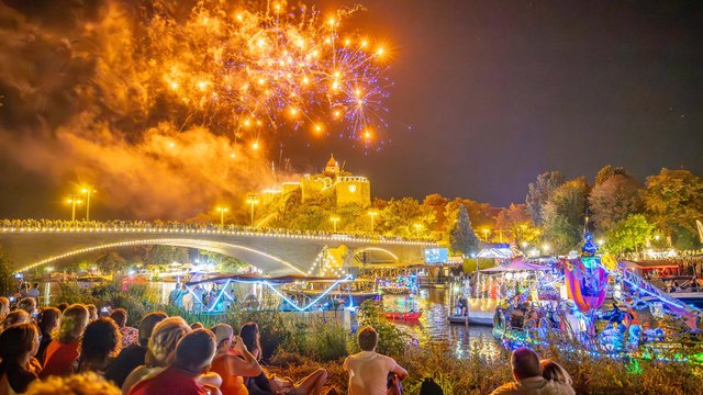 Großes Feuerwerk mit leuchtenden Farben und Explosionen am Nachthimmel über einem belebten Festplatz mit Menschen, die auf dem Boden sitzen und das Spektakel genießen. Bunte Lichterketten und Fahrgeschäfte im Hintergrund.