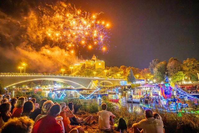 Thomas Ziegler, Stadt Halle (Saale) Großes Feuerwerk mit leuchtenden Farben und Explosionen am Nachthimmel über einem belebten Festplatz mit Menschen, die auf dem Boden sitzen und das Spektakel genießen. Bunte Lichterketten und Fahrgeschäfte im Hintergrund.