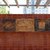 Mosaikwand mit drei Reliefs: links ein stilisiertes geometrisches Muster, in der Mitte eine liegende Person oder Figur in ovaler Vertiefung, rechts ein quadratisches Relief mit abstrakten Linien und Formen, eingebettet in rotbraune Kacheln.
