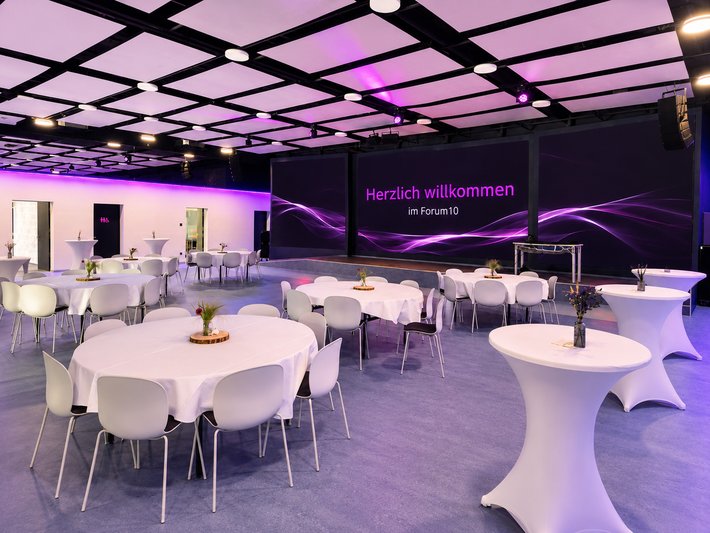 Großer, moderner Veranstaltungsraum mit mehreren runden weißen Tischen und Stühlen, die für eine Konferenz oder einen Empfang arrangiert sind. Im Hintergrund eine große Leinwand mit dem Begrüßungstext „Herzlich willkommen“ und lila Lichtakzenten.