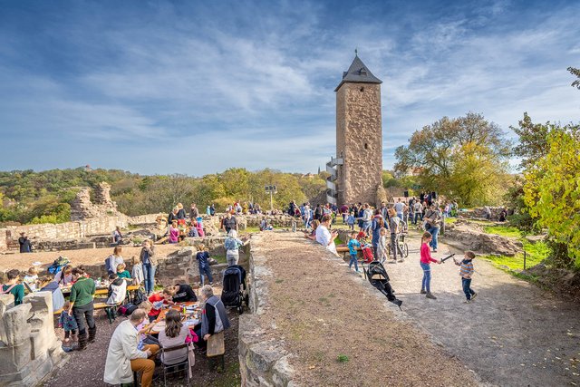 Große Menschenmenge auf einem Platz mit historischem Turm im Hintergrund, der an eine Burg erinnert. Viele Besucher sitzen auf Bänken oder stehen in Gruppen, bei sonnigem Wetter mit blauem Himmel und wenigen Wolken.