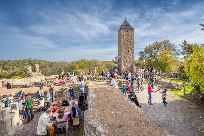 Große Menschenmenge auf einem Platz mit historischem Turm im Hintergrund, der an eine Burg erinnert. Viele Besucher sitzen auf Bänken oder stehen in Gruppen, bei sonnigem Wetter mit blauem Himmel und wenigen Wolken.