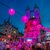 Abendliche Festbeleuchtung mit vielen bunten Laternen in Rosa und Violett vor einer historischen Kirche mit zwei Türmen, beleuchtet in Rot. Menschenmenge auf belebter Straße mit Ständen und festlicher Atmosphäre.