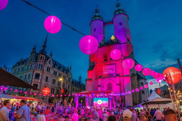 Abendliche Festbeleuchtung mit vielen pinken und violetten Luftlaternen über einem historischen Gebäude mit barocker Fassade, das an eine Kirche erinnert. Vor dem Gebäude ist ein belebter Platz mit vielen Menschen, die ein Fest oder eine Veranstaltung besuchen.