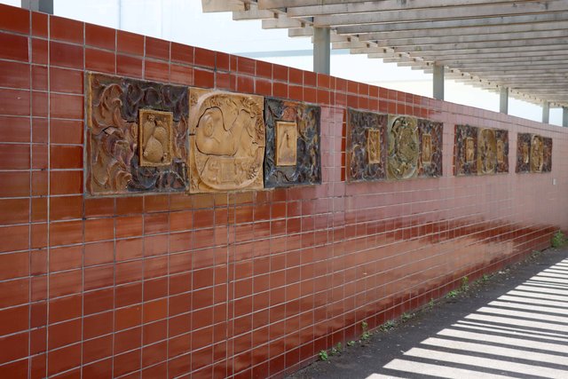 Eine Wand mit mehreren rechteckigen, farbenfrohen Mosaiktafeln, die historische oder kulturell bedeutende Porträts und Szenen darstellen, angeordnet in einer Reihe.