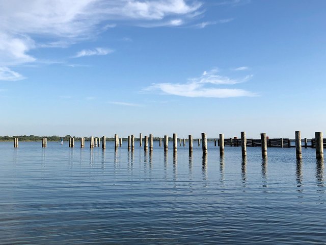Eine Reihe von Pfählen oder Pfahlreihen steht im ruhigen Wasser eines Sees oder einer Lagune, bei blauem Himmel mit wenigen Wolken.