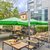 Ein gemütlicher Außenbereich mit mehreren Holztischen und Stühlen auf gepflastertem Boden. Große grüne Sonnenschirme bieten Schatten. Im Hintergrund moderne und historische Gebäude sowie grüne Büsche und Bäume.