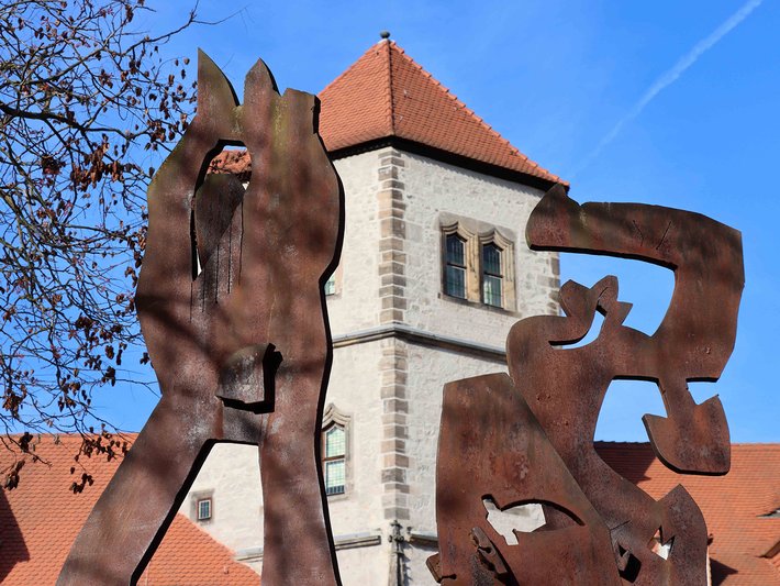 Große rostfarbene Skulptur vor einem historischen Gebäude mit Turm, die zwei sich umarmende oder kämpfende menschliche Figuren mit markanten, stilisierten Körperformen zeigt.