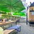 Moderner Außenbereich mit Holzbänken und Tischen unter großen grünen Sonnenschirmen. Im Hintergrund ein Holzpavillon mit Aussicht auf eine Stadt-Skyline. Pflanzkübel mit Grünpflanzen und gemütliche Sitzgelegenheiten auf gepflastertem Boden.