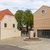 Zwei Gebäude in ländlichem Stil: ein helles, zweistöckiges Haus mit rotem Satteldach und ein modernes, einstufiges Gebäude mit flachem Dach und runder Fensteröffnung. Dazwischen ein runder Platz mit Pflasterung und zentralem runden Betonelement.