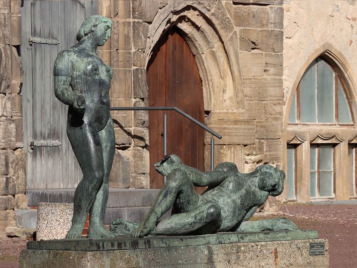 Bronzestatue eines stehenden Mannes, der einen liegenden, entspannten Hund mit den Händen stützt, vor historischer Backsteinfassade mit spitzbogigen Fenstern.