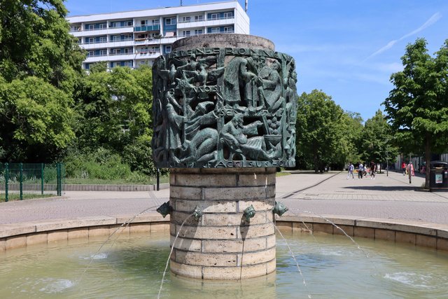 Bronzestatue mit mehreren nackten, sich umarmenden oder kämpfenden Figuren auf einem runden Sockel, umgeben von einem flachen Brunnenbecken mit klarem Wasser.