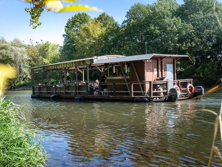 Ein gemütliches, großes Hausboot mit überdachtem Außenbereich und Sitzgelegenheiten fährt ruhig auf einem schmalen Fluss zwischen Bäumen und Grünflächen.