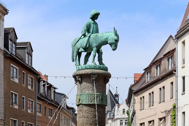 Bronzestatue eines reitenden Mannes auf einem Pferd, das auf einem steinernen Sockel in einer Stadtstraße steht, umgeben von historischen Gebäuden.