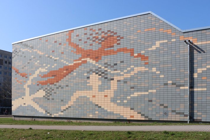 Großes Mosaik an einer Gebäudefassade, das eine springende oder laufende Figur in dynamischer Haltung zeigt, farblich dominiert von Orange und Grautönen auf hellem Hintergrund.