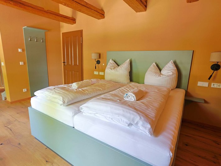 Gemütliches Hotelzimmer mit großem Bett, das mit weißer Bettwäsche und zwei Kissen in hellgrüner Farbe bezogen ist, dazu eine helle Decke. Zwei Nachttischlampen und ein Türrahmen mit Spiegel sind im Hintergrund sichtbar.