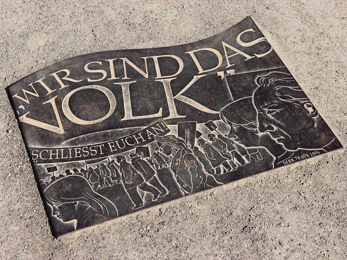 Ein Pflasterstein mit der Aufschrift „Wir sind das Volk“ und der Ergänzung „Schließt euch uns an“, kombiniert mit einer stilisierten Darstellung von Menschen, die sich verbinden. Symbolisiert friedliche Proteste und Solidarität.
