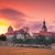 Abendliche Silhouette einer historischen Burg oder Festung mit roten Dächern und zwei markanten Türmen, umgeben von einer Mauer, bei Sonnenuntergang mit rötlich-orangefarbenem Himmel.