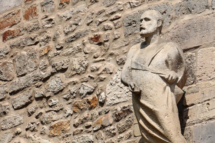Stehende Steinstatue eines Menschen mit langem Gewand, der eine Art Umhang oder Tuch über der Schulter trägt und eine Hand auf die Hüfte gestützt hat.