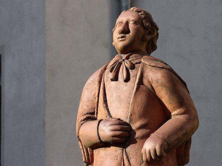Stehende Statue eines lächelnden Mannes in historischer Kleidung mit locker über der Brust gekreuzten Armen, aus hellem Stein gefertigt.