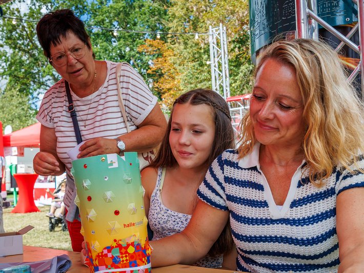 Drei Personen sitzen draußen an einem Tisch, eine ältere Frau und eine jüngere Frau helfen einem Mädchen, ein buntes, mit Süßigkeiten gefülltes Glas zu befüllen, vermutlich bei einer Feier oder einem Fest.