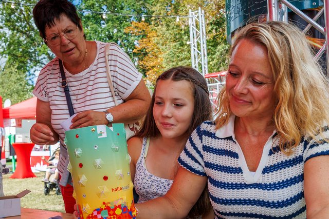 Drei Personen sitzen draußen an einem Tisch, eine ältere Frau und eine jüngere Frau helfen einem Mädchen, ein buntes, mit Süßigkeiten gefülltes Glas zu befüllen, vermutlich bei einer Feier oder einem Fest.
