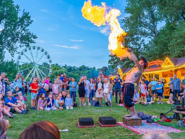 Ein Feuerkünstler entzündet mit einer Fackel eine gelbe Flamme, die er mit den Händen formt, während eine Gruppe von Zuschauern auf einer Wiese im Freien zusieht.