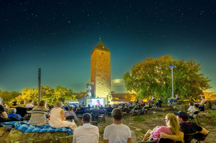 Abendveranstaltung im Freien mit vielen Zuschauern, die auf Liegestühlen und Decken sitzen und einem beleuchteten Turm im Hintergrund, der von Bäumen umgeben ist.