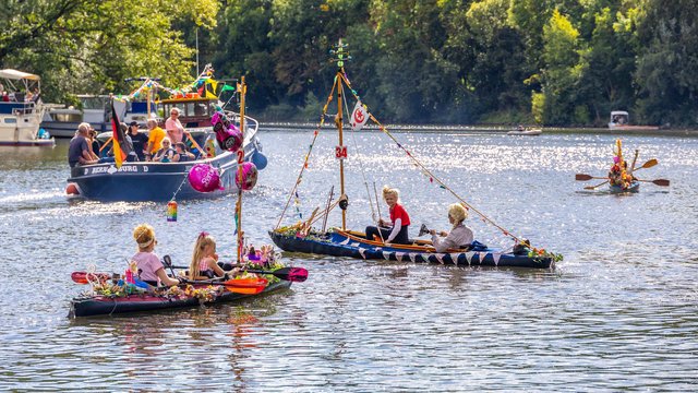 Bunte, selbstgebaute Floße mit Menschen darauf schwimmen auf einem See, umgeben von weiteren kleinen Booten und Kanus, bei einem fröhlichen Floß- oder Kanu-Wettbewerb.