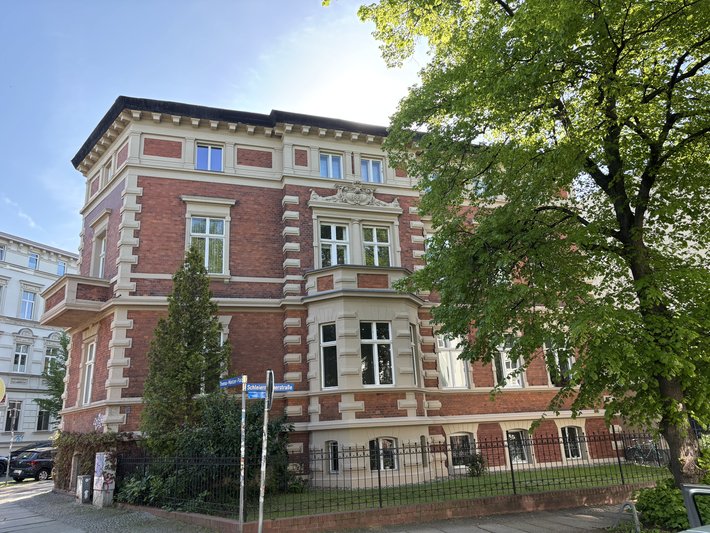 Historisches, dreistöckiges Backsteingebäude mit symmetrischer Fassade, zahlreichen großen Fenstern und Zierelementen wie Gesimsen und Pilastern. Eingebettet in eine grüne Umgebung mit Bäumen und einem gepflegten Vorgarten.