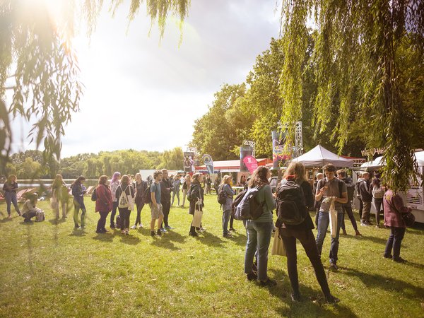Große Gruppe von Menschen auf einer Freiflächenveranstaltung mit Zelten und Ständen im Hintergrund, sonniges Wetter unter Bäumen.