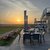 Sonnenuntergang über einer weiten Landschaft mit grasbewachsenem Feld und Start-/Landebahn im Hintergrund. Im Vordergrund mehrere Tische mit Stühlen auf einer Terrasse, teilweise mit Gläsern und Servietten bestückt.