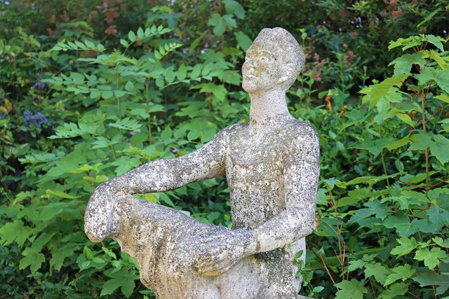 Skulptur einer sitzenden Person aus moosbewachsenem Material, die in meditativer Haltung mit geschlossenen Augen und gekreuzten Beinen auf einem Baumstumpf oder Sockel platziert ist, umgeben von grünem Pflanzenwerk.
