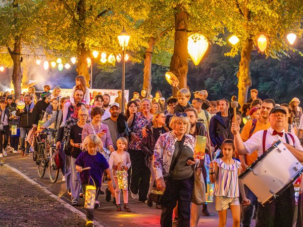 Große Gruppe von Menschen, darunter Familien mit Kindern, die bei einer abendlichen Veranstaltung mit Laternen und Lichterketten an Bäumen spazieren. Musikgruppe spielt im Hintergrund, fröhliche Stimmung.