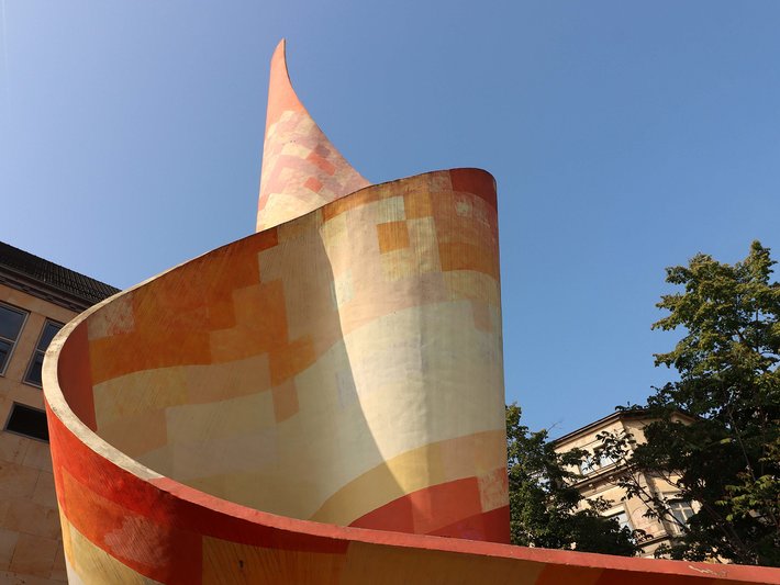 Moderne, wellenförmige Skulptur oder Architektur aus orange-beigen, geometrischen Platten, die sich nach oben verjüngt und an eine abstrakte, organische Form erinnert.