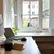 Arbeitsplatz mit Schreibtisch, auf dem ein Computerbildschirm, ein Laptop, eine Tastatur, eine Maus und ein Notizbuch liegen. Im Hintergrund ein Fenster mit Blick auf eine Straße und ein paar Menschen.