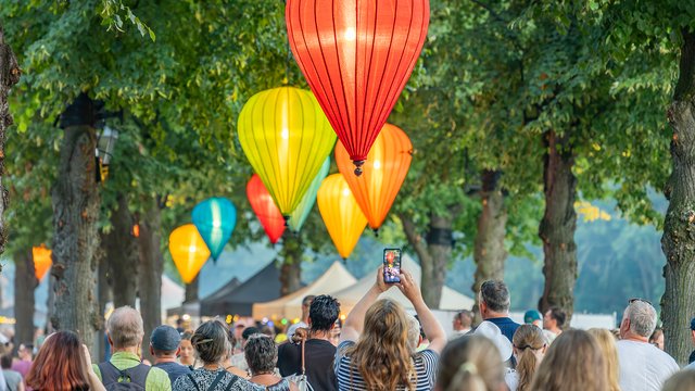 Eine Gruppe von Menschen beobachtet bunte Heißluftballons in verschiedenen Farben wie Rot, Orange und Gelb, die über einem Baum in der Luft schweben.