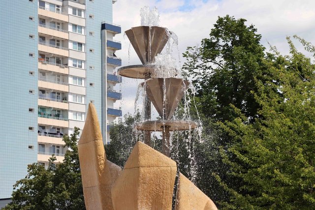 Skulptur mit goldener, trapezförmiger Basis und einer aufgesetzten, durchsichtigen Säule, aus der Wasser in einen kleinen Trichter fließt, umgeben von einem Brunnenbecken. Im Hintergrund ein Hochhaus und Bäume.