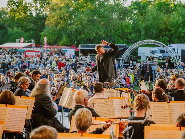 Großes Orchester spielt im Freien vor vollem Publikum, darunter viele Zuhörer auf Stühlen. Dirigent steht auf Podest und leitet mit erhobenen Armen. Musikinstrumente wie Streicher, Holz- und Blechbläser sind sichtbar.