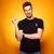 Ein Mann mit dunklem T-Shirt und Jeans steht lässig posierend, zeigt mit einer Hand ein Peace-Zeichen und hat die andere Armbeuge abgestützt.